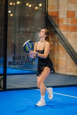 Riga, Letonya - 24 Ocak 2026: Padel turnuvası. Siyah elbiseli kadın padel oyuncu, mavi kortta cam duvarlı topa vurmaya hazırlanıyor.