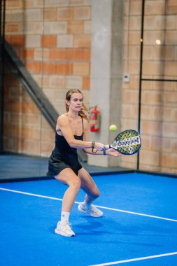 Riga, Letonya - 24 Ocak 2026: Padel turnuvası. Siyah elbiseli kadın padel oyuncu mavi kortta duvarları görünen bir topa vurabilir.