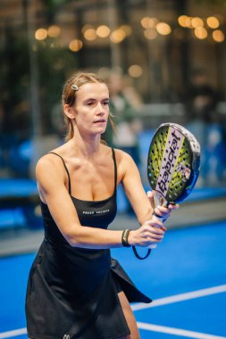 Riga, Letonya - 24 Ocak 2026: Padel turnuvası. Siyah elbiseli kadın padel oyuncu, arka planı bulanık mavi bir kortta yeşil ve siyah raket tutuyor.