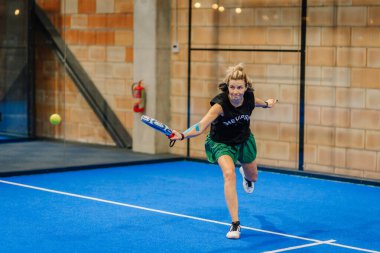 Riga, Letonya - 24 Ocak 2026: Padel turnuvası. Siyah tişörtlü ve yeşil şortlu bayan padel oyuncu, mavi kortta camdan duvarları olan bir topa vurmak için öne atlıyor.