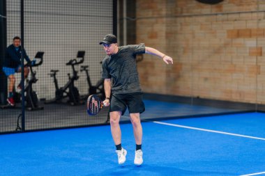 Riga, Letonya - 24 Ocak 2026: Padel turnuvası. Siyah elbiseli erkek sporcu, arka planda spor bisikletleriyle mavi kortta Padel oynuyor.