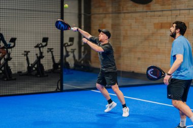 Riga, Letonya - 24 Ocak 2026: Padel turnuvası. Biri siyah tişörtlü ve şapkalı iki adam içeride Padel oynuyor. Topa vurmaya hazırlanıyor, diğeri mavi gömlekli.