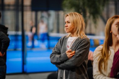 Riga, Letonya - 24 Ocak 2026: Padel turnuvası. Gri hırkalı sarışın kadın, mavi döşemeli bir spor tesisinde sahada Padel oyuncularını izliyor.