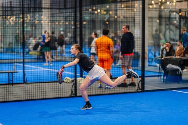 Riga, Letonya - 24 Ocak 2026: Padel turnuvası. Siyah şortlu ve beyaz şortlu bayan sporcu, mavi sahada seyircilerin görebileceği bir top servis ediyor.