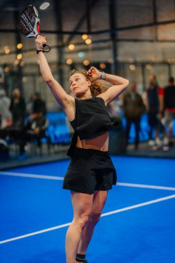 Riga, Letonya - 24 Ocak 2026: Padel turnuvası. Siyah spor giyimli bayan oyuncu, mavi sahada seyircilerle yapılacak bir Padel maçına hazırlanıyor.