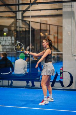 Riga, Letonya - 24 Ocak 2026: Padel turnuvası. Gri etekli kadın padel oyuncu, arkasında seyirciler varken mavi sahada topa vuracak pozisyonda.