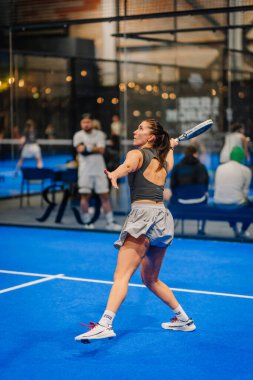 Riga, Letonya - 24 Ocak 2026: Padel turnuvası. Gri elbiseli bayan padel oyuncu, mavi sahada topa vurmaya hazırlanıyor.