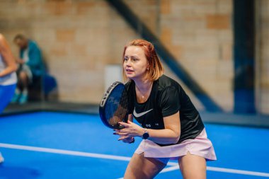 Riga, Letonya - 24 Ocak 2026: Padel turnuvası. Siyah tişörtlü ve pembe etekli bayan padel oyuncu mavi kortta başka bir oyuncuyla servis yapmaya hazırlanıyor.