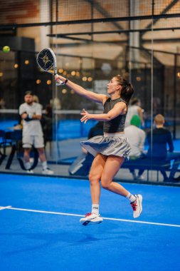 Riga, Letonya - 24 Ocak 2026: Padel turnuvası. Gri etekli ve siyah bluzlu bayan padel oyuncu, mavi sahada seyircilerin gözü önünde topa vurmaya hazırlanıyor.