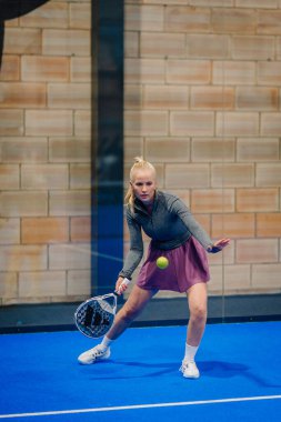 Riga, Letonya - 24 Ocak 2026: Padel turnuvası. Gri bluzlu ve pembe etekli bayan padel oyuncu mavi kortta tuğla duvarla topa vuracak pozisyonda.