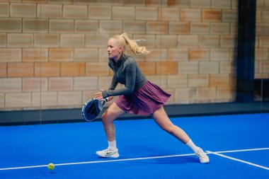 Riga, Letonya - 24 Ocak 2026: Padel turnuvası. Gri uzun kollu bluz ve pembe etekli bayan padel oyuncu mavi kortta topa vurmaya hazır.