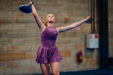 Riga, Letonya - 24 Ocak 2026: Padel turnuvası. Mor spor giyim giyen padel sporcusu arka tarafta duvar ve elektrik paneli olan iç sahada hizmet vermeye hazırlanıyor.