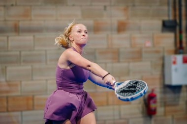Riga, Letonya - 24 Ocak 2026: Padel turnuvası. Mor elbiseli kadın padel oyuncu, duvarlı bir evde oynarken raket sallıyor.