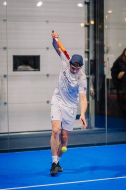 Riga, Letonya - 24 Ocak 2026: Padel turnuvası. Beyaz elbiseli erkek padel oyuncusu mavi bir sahada cam duvarları ve seyircileri olan bir topa hizmet etmeye hazırlanıyor.