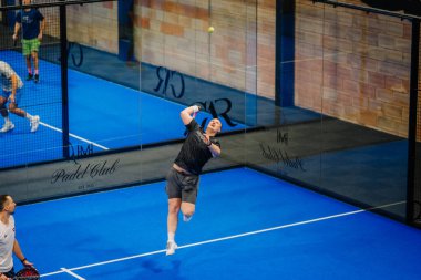 Riga, Letonya - 24 Ocak 2026: Padel turnuvası. Siyah gömlekli erkek sporcu, mavi zemin ve cam duvarların yer aldığı kapalı bir padel sahada top servis ediyor.