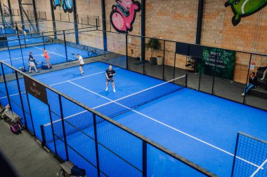 Riga, Letonya - 24 Ocak 2026: Padel turnuvası. Padel oyuncuları, renkli duvar sanatı ve eskrim sanatının yer aldığı bir spor tesisinde mavi sahada yarışıyorlar.