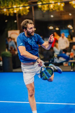 Riga, Letonya - 24 Ocak 2026: Padel turnuvası. Mavi tişörtlü ve beyaz şortlu erkek padel oyuncu, mavi sahada seyircilerin gözü önünde servis yapmaya hazırlanıyor.