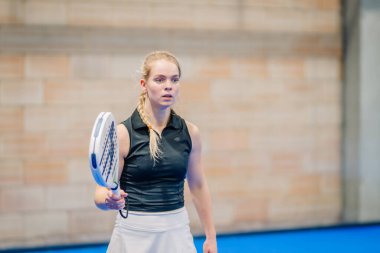 Riga, Letonya - 24 Ocak 2026: Padel turnuvası. Siyah ve beyaz etekli bayan padel oyuncu mavi bir kortta tuğladan bir duvarla hizmet etmeye hazır.