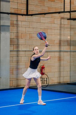 Riga, Letonya - 24 Ocak 2026: Padel turnuvası. Siyah ve beyaz etekli kadın padel atlet, kapalı alanda mavi bir kortta topa vurmaya hazırlanıyor.