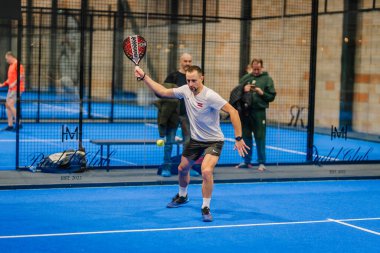 Riga, Letonya - 24 Ocak 2026: Padel turnuvası. Beyaz tişörtlü ve şortlu bir erkek sporcu, kapalı alanda mavi bir kortta topa vuruyor.