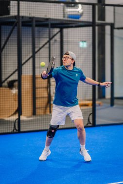 Riga, Letonya - 24 Ocak 2026: Padel turnuvası. Mavi tişörtlü ve şortlu erkek padel oyuncusu mavi bir kortta topa ağla vurmaya hazırlanıyor.
