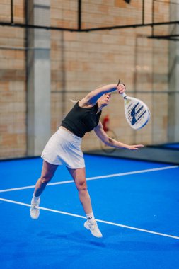 Riga, Letonya - 24 Ocak 2026: Padel turnuvası. Siyah atlet ve beyaz etekli kadın padel atlet mavi kortta ağlı bir topla servis yapıyor.