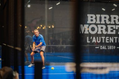 Riga, Letonya - 24 Ocak 2026: Padel turnuvası. Mavi tişörtlü erkek padel oyuncu duvarında damgalamayla iç sahada topa vurmaya hazır.