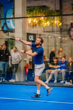 Riga, Letonya - 24 Ocak 2026: Padel turnuvası. Mavi tişörtlü erkek sporcu, seyircilerle birlikte kapalı bir sahada bir padel topu servis etmeye hazırlanıyor.