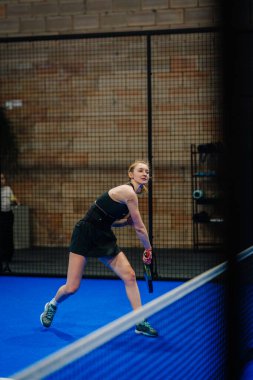 Riga, Letonya - 24 Ocak 2026: Padel turnuvası. Siyah elbiseli bayan sporcu, mavi bir padel kortunda topa vurmaya hazırlanıyor.