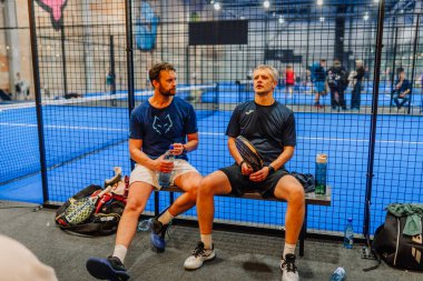 Riga, Letonya - 24 Ocak 2026: Padel turnuvası. Atletik giyimli iki adam padel kortunda bir bankta oturuyor, ellerinde mavi bir kortla su şişeleri tutuyorlar.