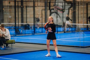 Riga, Letonya - 24 Ocak 2026: Padel turnuvası. Siyah elbiseli kadın padel oyuncu, arka planda izleyicilerin olduğu bir raket tutarak mavi bir kortta duruyor.