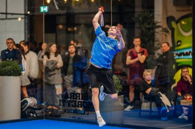 Riga, Letonya - 24 Ocak 2026: Padel turnuvası. Mavi tişörtlü erkek padel oyuncu, arka planda seyircilerin olduğu kapalı bir sahada topa vurmak için sıçrıyor.