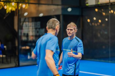 Riga, Letonya - 24 Ocak 2026: Padel turnuvası. Mavi atletizm tişörtlü iki adam mavi padel kortunda cam duvarlar ve iç aydınlatma ile sohbet ediyorlar.