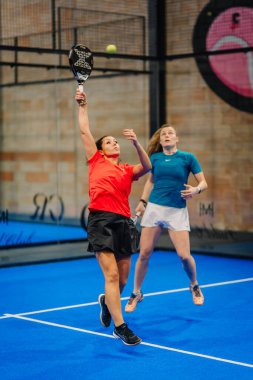 Riga, Letonya - 24 Ocak 2026: Padel turnuvası. İki bayan oyuncu kapalı alanda mavi bir sahada maç yapıyor, biri servis atıyor ve diğeri cevap vermeye hazır.