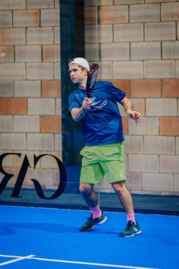 Riga, Letonya - 24 Ocak 2026: Padel turnuvası. Mavi ve yeşil giyimli erkek padel oyuncusu, mavi sahada cam panellerle topa vurmak için bizzat kendisi pozisyon aldı.