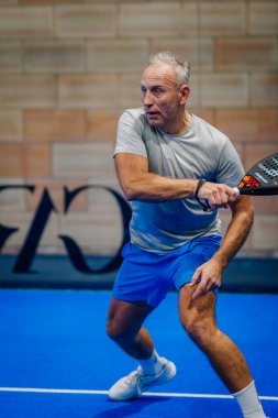 Riga, Letonya - 24 Ocak 2026: Padel turnuvası. Gri tişörtlü ve mavi şortlu son sınıf erkek oyuncu mavi bir padel kortta, raketle topa vurmaya hazır.