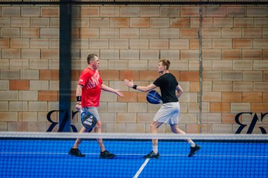 Riga, Letonya - 24 Ocak 2026: Padel turnuvası. Spor kıyafetli iki adam, modern bir bina içinde mavi bir padel kortunda birbirlerini selamlıyorlar.