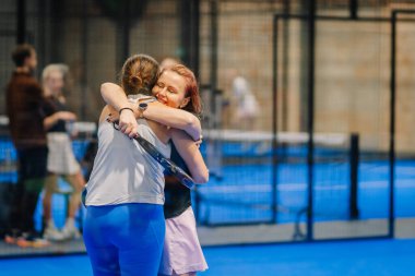 Riga, Letonya - 24 Ocak 2026: Padel turnuvası. Spor kıyafetli iki bayan oyuncu, arka planda eskrim ve diğer oyuncularla mavi bir padel kortunda sarılıyorlar.