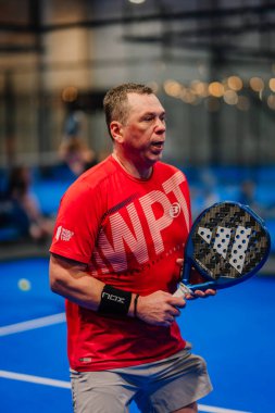 Riga, Letonya - 24 Ocak 2026: Padel turnuvası. Kırmızı tişört ve bileklik giyen erkek padel oyuncusu mavi kortta topa vurmaya hazırlanıyor.