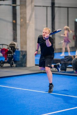 Riga, Letonya - 24 Ocak 2026: Padel turnuvası. Siyah tişörtlü ve şortlu bir erkek atlet, mavi bir sahada seyircilerle birlikte bir padel topu servis ediyor.