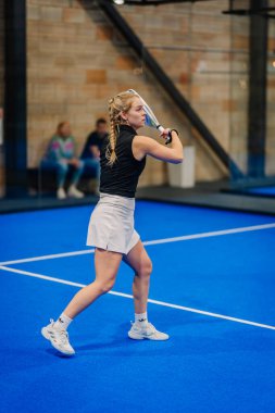 Riga, Letonya - 24 Ocak 2026: Padel turnuvası. Siyah tişörtlü ve beyaz etekli bayan padel oyuncu mavi bir kortta raket sallıyor.