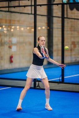 Riga, Letonya - 24 Ocak 2026: Padel turnuvası. Siyah bluzlu ve beyaz etekli kadın padel oyuncu mavi kortta topa vurmaya hazırlanıyor.