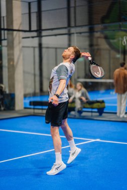 Riga, Letonya - 24 Ocak 2026: Padel turnuvası. Spor giyim sektöründe erkek atlet, arka planda oyuncular ve izleyicilerle mavi sahada bir padel topu sunuyor.