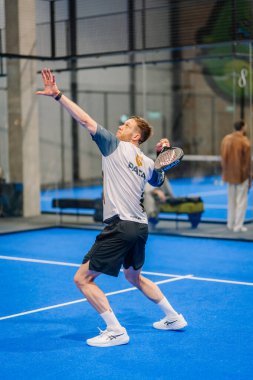 Riga, Letonya - 24 Ocak 2026: Padel turnuvası. Erkek sporcu, mavi bir sahada spor kıyafetleri içinde iç mekan logosu olan padel topu servis ediyor.