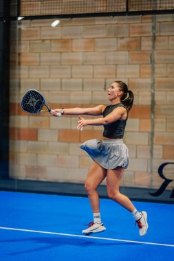 Riga, Letonya - 24 Ocak 2026: Padel turnuvası. Siyah kolsuz üst ve gri etekli bayan padel oyuncu mavi kortta topa vurmaya hazırlanıyor.