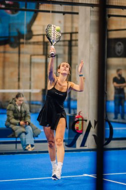 Riga, Letonya - 24 Ocak 2026: Padel turnuvası. Siyah elbiseli bayan padel oyuncu, arka planda seyircilerin olduğu mavi bir sahada topa hizmet etmeye hazırlanıyor.