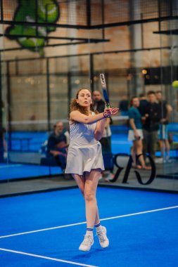 Riga, Letonya - 24 Ocak 2026: Padel turnuvası. Gri elbiseli bayan padel oyuncu, arka planda seyircilerin olduğu mavi bir sahada topa vurmaya hazırlanıyor.