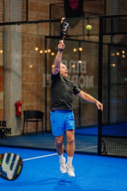 Riga, Letonya - 24 Ocak 2026: Padel turnuvası. Siyah tişörtlü ve mavi şortlu erkek padel oyuncusu şık bir tasarıma sahip bir iç sahada top servis ediyor.