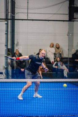 Riga, Letonya - 24 Ocak 2026: Padel turnuvası. Siyah tişörtlü ve şortlu bir erkek sporcu mavi bir padel kortunda topa vuruyor ve seyirciler izliyor.