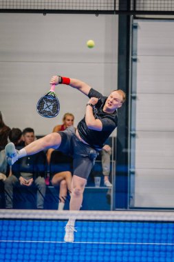 Riga, Letonya - 24 Ocak 2026: Padel turnuvası. Siyah tişörtlü ve şortlu bir erkek atlet, kapalı sahada seyirciler izlerken padel topu servis ediyor.